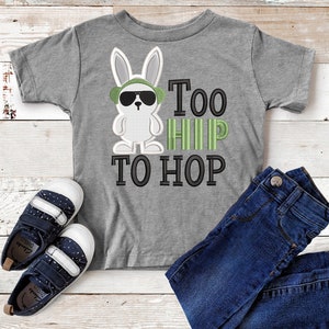 Pode incluir: Uma camiseta cinza com um coelho de desenho animado usando óculos de sol e um chapéu verde. O texto "Too Hip To Hop" é impresso em preto e verde. A camiseta é combinada com jeans azul e sapatos azul marinho.
