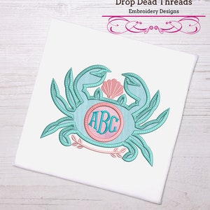 Könnte beinhalten: Eine gestickte Darstellung einer blauen Krabbe mit einer rosa Muschel und einem Kreis mit den Buchstaben ABC darin. Das Design ist auf weißem Hintergrund.