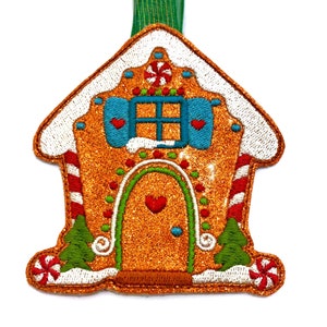 Peut inclure: Une décoration de maison en pain d'épices avec du glaçage blanc, des cannes en sucre rouge et des sapins verts. La maison est décorée d'une fenêtre bleue et d'un cœur rouge sur la porte.