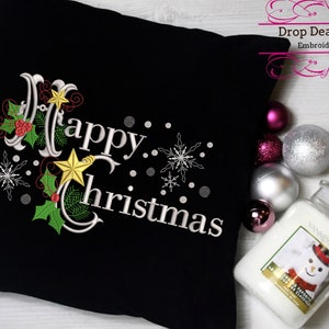 May include: A black pillow with white embroidery that reads "Happy Christmas". The embroidery includes holly, stars, and snowflakes. The pillow is surrounded by silver and pink ornaments and a white candle.
