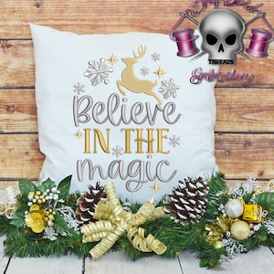 Könnte beinhalten: Ein weißes Kissen mit goldener und silberner Stickerei, auf dem "Glaube an die Magie" steht. Die Stickerei enthält ein Rentier, Schneeflocken und Sterne. Das Kissen ist von einem grünen Kranz mit goldener Schleife, Tannenzapfen und goldenen Ornamenten umgeben.