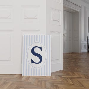 Puede incluir: Un cuadro con la letra "S" en azul marino, dentro de un óvalo sobre un fondo a rayas azules y blancas. La impresión se muestra sobre un suelo de madera, apoyada contra una puerta blanca. Adecuado para una habitación infantil.