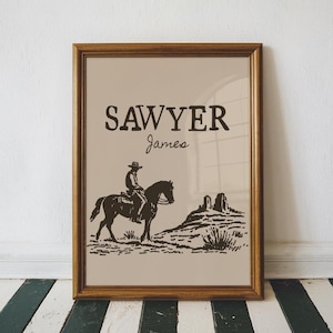 May include: A framed sepia-toned print showing a cowboy on horseback, with the name "SAWYER" above and "James" below in a script font. The image depicts a desert scene with a mountain and sparse vegetation.