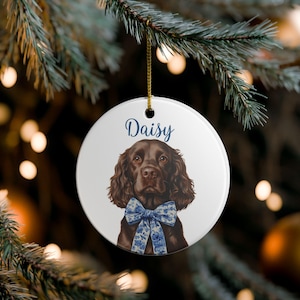May include: A round, white Christmas ornament featuring a brown dog with a blue floral bow tie. The word "Daisy" is written in blue script above the dog's head. The ornament hangs from a gold string against a blurred background of Christmas lights.