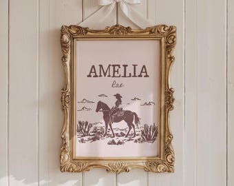 May include: A framed print featuring a cowgirl on horseback in a desert landscape. The print is in a gold ornate frame and is suspended by a white ribbon and bow. The name "AMELIA Rae" is printed at the top of the image.