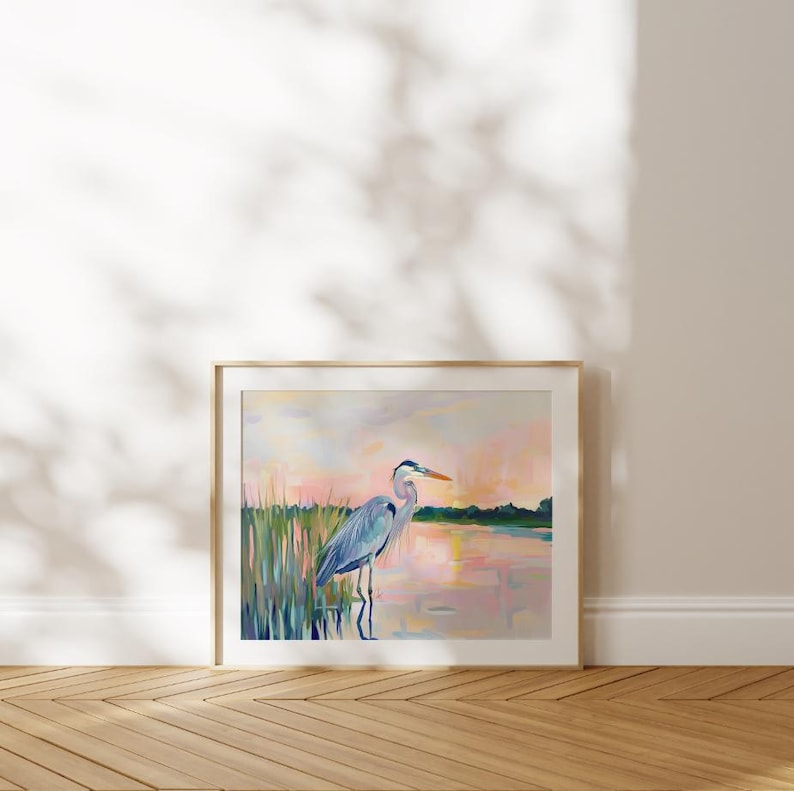 May include: A painting of a blue heron standing in a marsh at sunset. The heron is facing to the right and has its head turned to the left. The water is a light pink color and the sky is a light blue color. The marsh is a dark green color.