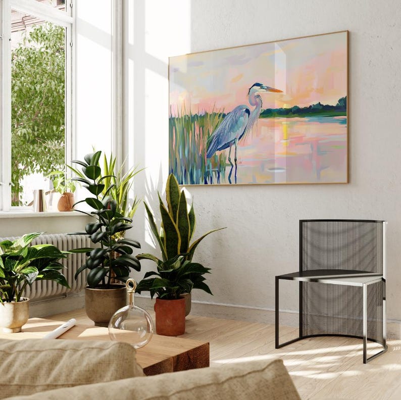 May include: A painting of a blue heron standing in a lake at sunset. The painting is framed in gold and hanging on a white wall. A black metal chair is in the foreground.