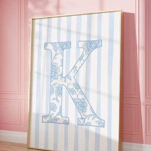 Op de afbeelding: Een ingelijste verticale wandkunstprint met een grote hoofdletter K in een lichtblauw bloemenpatroon. De letter is gecentreerd tegen een achtergrond van verticale lichtblauwe en witte strepen, alles in een dunne gouden lijst.