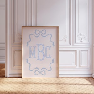 Op de afbeelding: Een ingelijste print met een lichtroze achtergrond, met de letters "MBC" in een lichtblauw bloemmotief. Lichtblauwe linten en strikken omringen de letters. De lijst is lichtbruin.