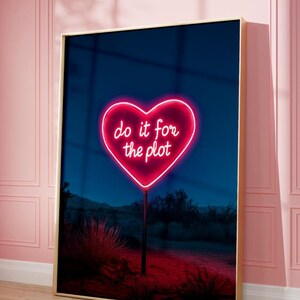 Puede incluir: Una impresión enmarcada de un letrero de neón en forma de corazón que dice "do it for the plot" en un paisaje desértico de noche.