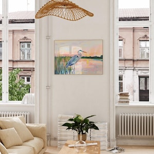 May include: A painting of a blue heron standing in a marsh at sunset. The heron is facing right and has its head turned to the left. The water is a light pink color and the sky is a light orange color.