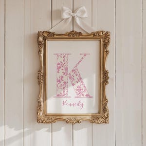 May include: A framed print featuring the letter "K" in a floral pattern, with the name "Kennedy" below. The print is in a gold ornate frame and is suspended by a white ribbon and bow against a white wooden background.
