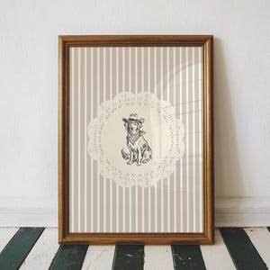 Könnte beinhalten: Gerahmter Druck mit einer Hundeillustration mit Cowboyhut und Halstuch. Der Hund befindet sich auf einer cremefarbenen Spitze vor einem gestreiften Hintergrund in Beige und Weiß. Der Rahmen hat eine warme braune Farbe.