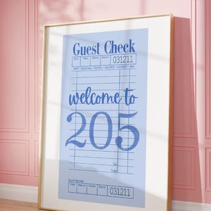 May include: A framed vertical print featuring a light blue restaurant guest check design. The text reads Guest Check, welcome to, and 205, with fields for date, table, guests, server, tax, and total. The print is enclosed in a thin light wood frame.