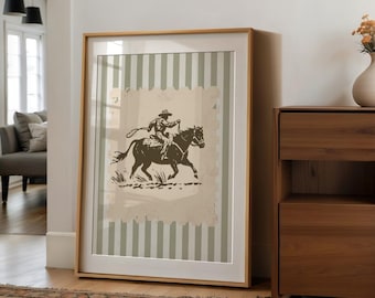 May include: Framed artwork featuring a cowboy riding a horse, rendered in a vintage style. The image is set against a backdrop of vertical stripes in muted green and white. The frame is a light wood color, adding a rustic touch.