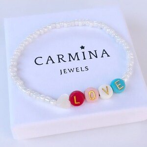 Könnte beinhalten: Weißes Perlenarmband mit dem Wort "LOVE" in bunten Perlen mit goldenen Buchstaben. Das Armband befindet sich in einer weißen Box mit dem Text "CARMINA JEWELS" darauf.
