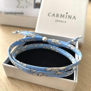 Könnte beinhalten: Ein hellblaues Wickelarmband mit weißem Blumenmuster, präsentiert in einer offenen weißen Schachtel. Auf der Schachtel steht der Markenname "CARMINA JEWELS". Das Armband ist um einen schwarzen Einsatz gewickelt. Ein zartes Schmuckstück.