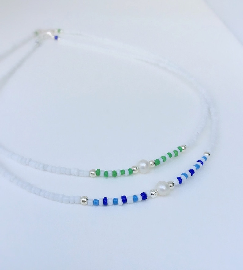 May include: A delicate white beaded necklace with two strands of colourful beads. One strand features blue and white beads, while the other strand features green and white beads. Both strands are accented with a single white pearl.