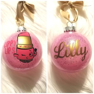 May include: Two pink glitter Christmas ornaments with gold accents. One ornament features a cartoon lipstick character with the text "Lippy Lips". The other ornament has the name "Lilly" written in gold.