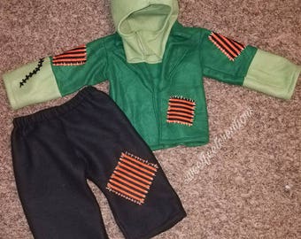 May include: Green and black fleece Frankenstein costume for a child. The costume includes a hooded jacket with stitched seams and orange and black patches, and black fleece trousers with orange and black patches.