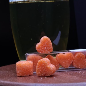 Peut inclure: Un verre de champagne posé sur une surface en bois à côté d'un récipient en plastique transparent rempli de bonbons en forme de cœur orange.