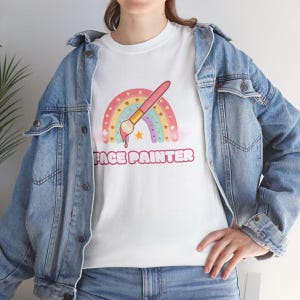 Op de afbeelding: Wit T-shirt met een regenboog- en penseelontwerp. De tekst "FACE PAINTER" is onder het ontwerp gedrukt.