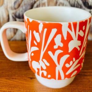 May include: A ceramic coffee mug with a white handle and base. The mug's exterior is bright orange with a white floral pattern. The interior of the mug is white. The mug is sitting on a wooden surface.