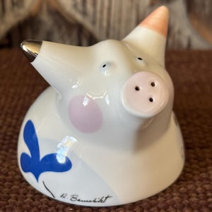 May include: A white ceramic pig-shaped salt shaker with a silver snout and pink ear. The pig has a light purple cheek and blue floral design. The artist's signature, "R. Baumlikt", is written on the bottom.