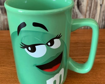 May include: Green ceramic coffee mug with a cartoon face of a green M&M's character. The character has a winking eye, eyelashes, and a big smile.