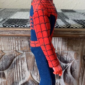 K&ouml;nnte beinhalten: Eine Vintage-Spider-Man-Actionfigur. Die Figur ist haupts&auml;chlich rot und blau, mit einer roten Maske und einem blauen K&ouml;rper. Das Kost&uuml;m hat ein Spinnennetz-Design. Die Figur steht auf einer Holzoberfl&auml;che.