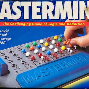 May include: Mastermind board game box featuring the title in large white letters on a blue background. The game board is dark gray with rows of holes and colorful plastic pegs in red, blue, yellow, white, and green. The box includes text.