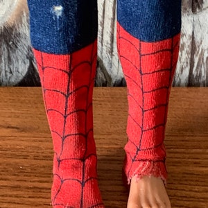 K&ouml;nnte beinhalten: Nahaufnahme der Beine einer Puppe, die blauen, denimartigen Stoff und rote Socken mit einem schwarzen Spinnennetz-Design tr&auml;gt. Die Socken sind an den Zehen zerrissen. Die F&uuml;&szlig;e der Puppe sind sichtbar.