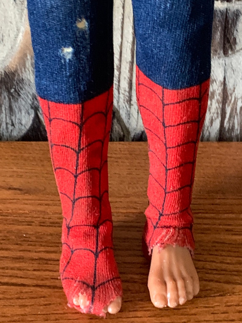 K&ouml;nnte beinhalten: Nahaufnahme der Beine einer Puppe, die blauen, denimartigen Stoff und rote Socken mit einem schwarzen Spinnennetz-Design tr&auml;gt. Die Socken sind an den Zehen zerrissen. Die F&uuml;&szlig;e der Puppe sind sichtbar.
