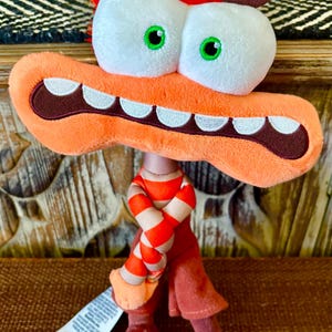May include: A plush toy featuring a large orange head with wide white eyes, green pupils, and a broad mouth showing white teeth. The character has messy orange hair, brown eyebrows, and wears a red and orange striped shirt with dark red pants and shoes.