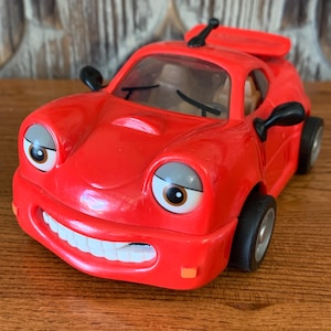 May include: A red toy car with a smiling face, large eyes, and a black rearview mirror. The car has a black spoiler on the back.