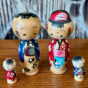 May include: Four wooden Japanese Kokeshi dolls. Two larger dolls have black hair and are wearing blue and red patterned kimonos. Two smaller dolls are red and blue. The dolls are on a wooden surface.
