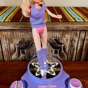 May include: A vintage Dream Dancer doll in a purple leotard, pink briefs, and leg warmers, with a matching headband. The doll is on a purple base with the words "Dream Dancer" and buttons labeled "Pose" and "Ballet".