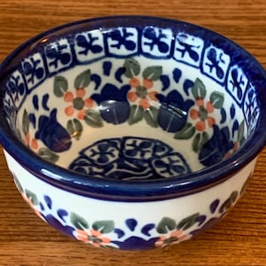 May include: A small, white ceramic bowl with a blue rim and a floral pattern in shades of blue, green, and pink.