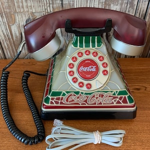 May include: A vintage-style rotary phone with a stained glass design featuring red, green, and white colors. The phone has a Coca-Cola logo and the words "Coca-Cola" on the base. The phone has a black coiled cord and a gray telephone cord.