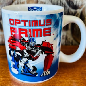 May include: A white ceramic mug featuring a graphic of the character Optimus Prime in red, blue, and silver. The text "OPTIMUS PRIME" is printed in bold red letters above the character. A blue Autobot logo is visible on the inner rim of the mug.