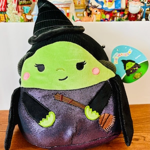 May include: A plush Wicked Witch Squishmallow with a green face, black hat, and purple dress. The witch has pink cheeks, black eyes, and a small smile. She holds a brown broom and has a tag that says "Original Squishmallows".