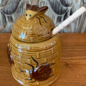 May include: A ceramic honey pot shaped like a beehive with a bee on top and a wooden honey dipper. The pot is decorated with brown and yellow glaze and has a beehive pattern.