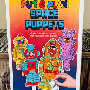 May include: A vintage Sesame Street "Put & Play Space Puppets" activity set. The box features colorful illustrations of space-themed puppets, including a robot and characters in astronaut suits. The text reads "dress four stick puppets with reusable vinyl pieces."