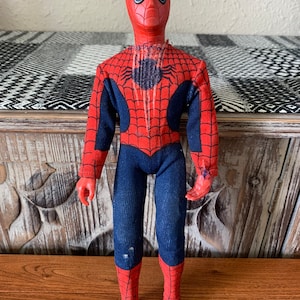 K&ouml;nnte beinhalten: Eine Vintage-Spider-Man-Actionfigur. Die Figur tr&auml;gt ein rot-blaues Kost&uuml;m mit Spinnennetz-Design. Der Kopf ist rot mit wei&szlig;en Augenlinsen. Die Figur steht auf einer Holzoberfl&auml;che.