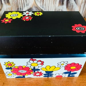May include: A black metal box with a floral design. The top of the box is black with yellow and red flowers. The bottom of the box is white with yellow, red, and white flowers.