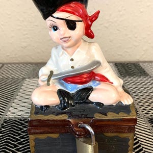 May include: A ceramic pirate figurine with a black hat, red bandana, and an eyepatch sits on top of a locked wooden treasure chest. The chest has a brown lid with gold accents and a black base.