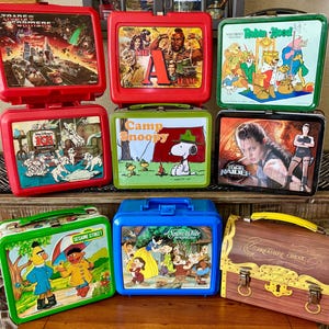 May include: A collection of eight rectangular metal and plastic lunch boxes featuring various pop culture themes including Transformers, The A-Team, Robin Hood, 101 Dalmatians, Camp Snoopy, Tomb Raider, Sesame Street, Snow White, and a treasure chest design.