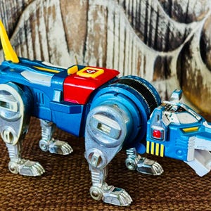 May include: A blue robotic lion toy featuring silver legs, a yellow tail, and a red square panel on its back marked with the number 4. The head includes a red eye detail, yellow accents, and a white jaw section, with mechanical detailing throughout.