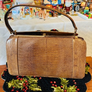 May include: A vintage brown handbag with a textured, reptile-like pattern. The rectangular bag has a curved handle and a gold-toned clasp. It rests on a black, scalloped mat decorated with holly and red berries, suggesting a holiday theme.