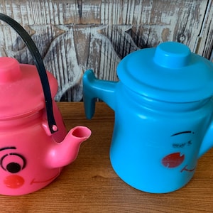 May include: Two plastic toy teapots with painted faces. One is pink with a black handle and the other is blue with a white handle.